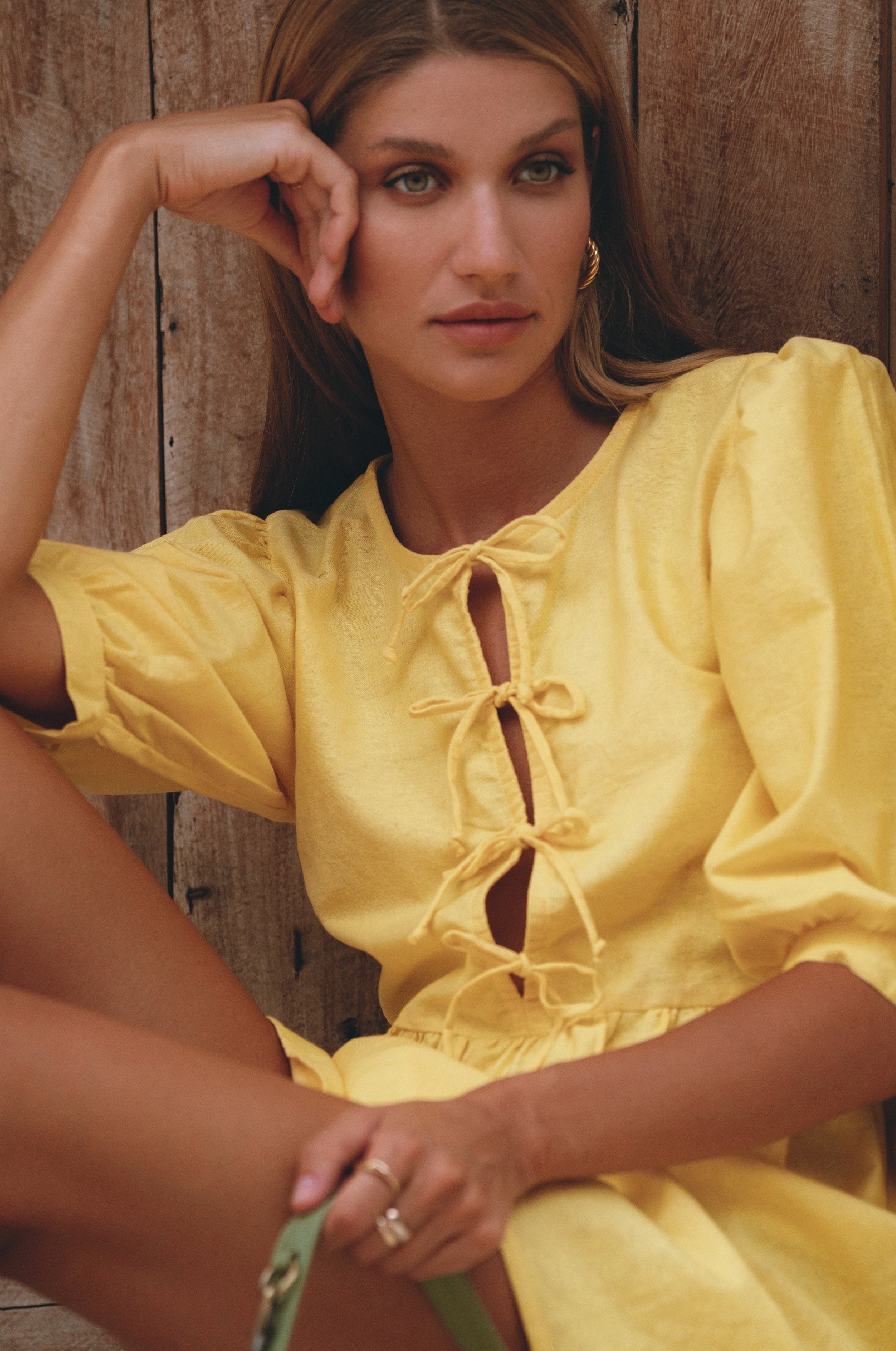 Woman wearing a yellow linen mini dress with puff sleeves and ties in the front bodice, leaning against a wood wall, front view closeup