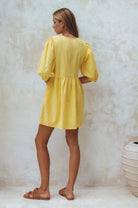 Woman wearing a yellow linen mini dress with puff sleeves standing against a textured white wall, back view
