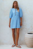 Woman wearing a light blue linen mini dress with puff sleeves standing against a textured white wall, front view
