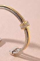 A close-up of a cable-style cuff bracelet featuring twisted silver-toned and yellow-gold metal bands, with a decorative clasp set with small clear stones, photographed on a soft beige background.