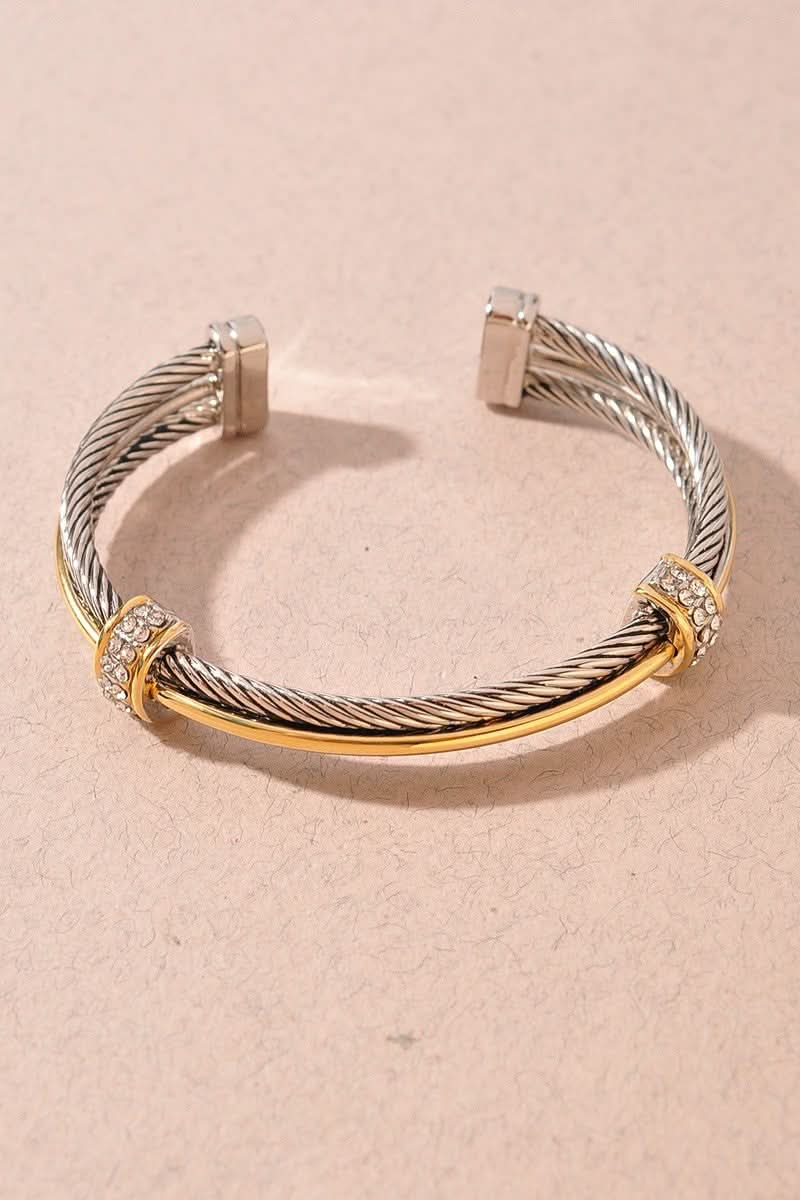 A cable-style cuff bracelet with a twisted silver-tone band, a polished gold-tone center bar, and small clear stone accents on the gold ends, photographed on a warm beige background.
