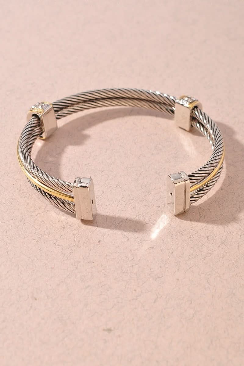 A silver-toned cable cuff bracelet with polished rectangular silver end pieces and small gold-toned accents, photographed on a soft pink background.