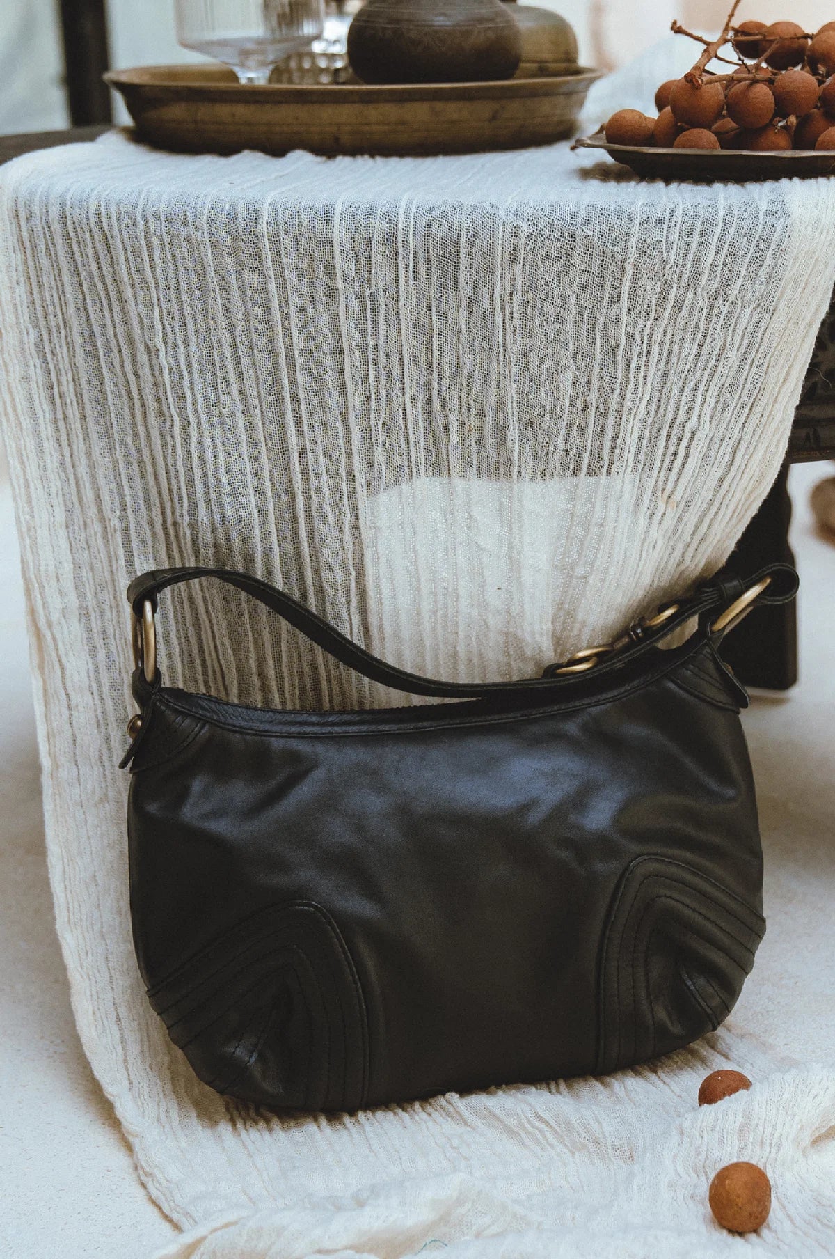A black leather shoulder bag with gold-tone hardware, resting on a draped white textured fabric next to a table with decorative fruit.