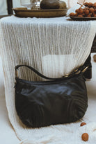 A black leather shoulder bag with gold-tone hardware, resting on a draped white textured fabric next to a table with decorative fruit.