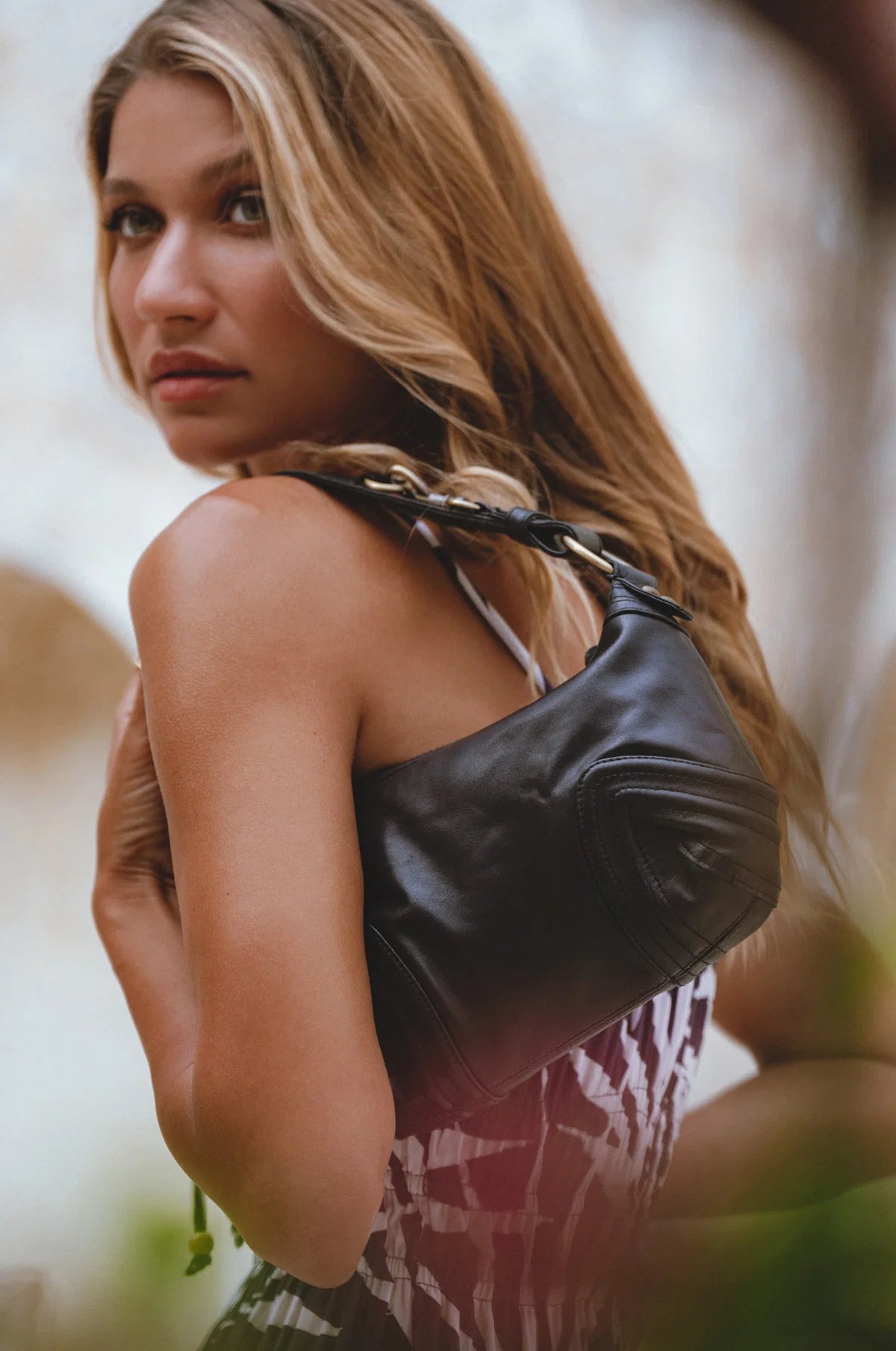 A woman with long blonde hair is shown from the back wearing a black leather shoulder bag with a braided strap over a patterned top, set against a soft-focus outdoor background.