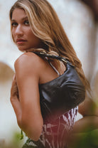 A woman with long blonde hair is shown from the back wearing a black leather shoulder bag with a braided strap over a patterned top, set against a soft-focus outdoor background.