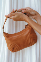 Tan leather handbag held by a person against a white curtain background 