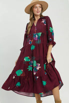 A woman modeling a burgundy midi dress with a colorful floral print, paired with a wide-brimmed straw hat and ankle boots against a plain light background.