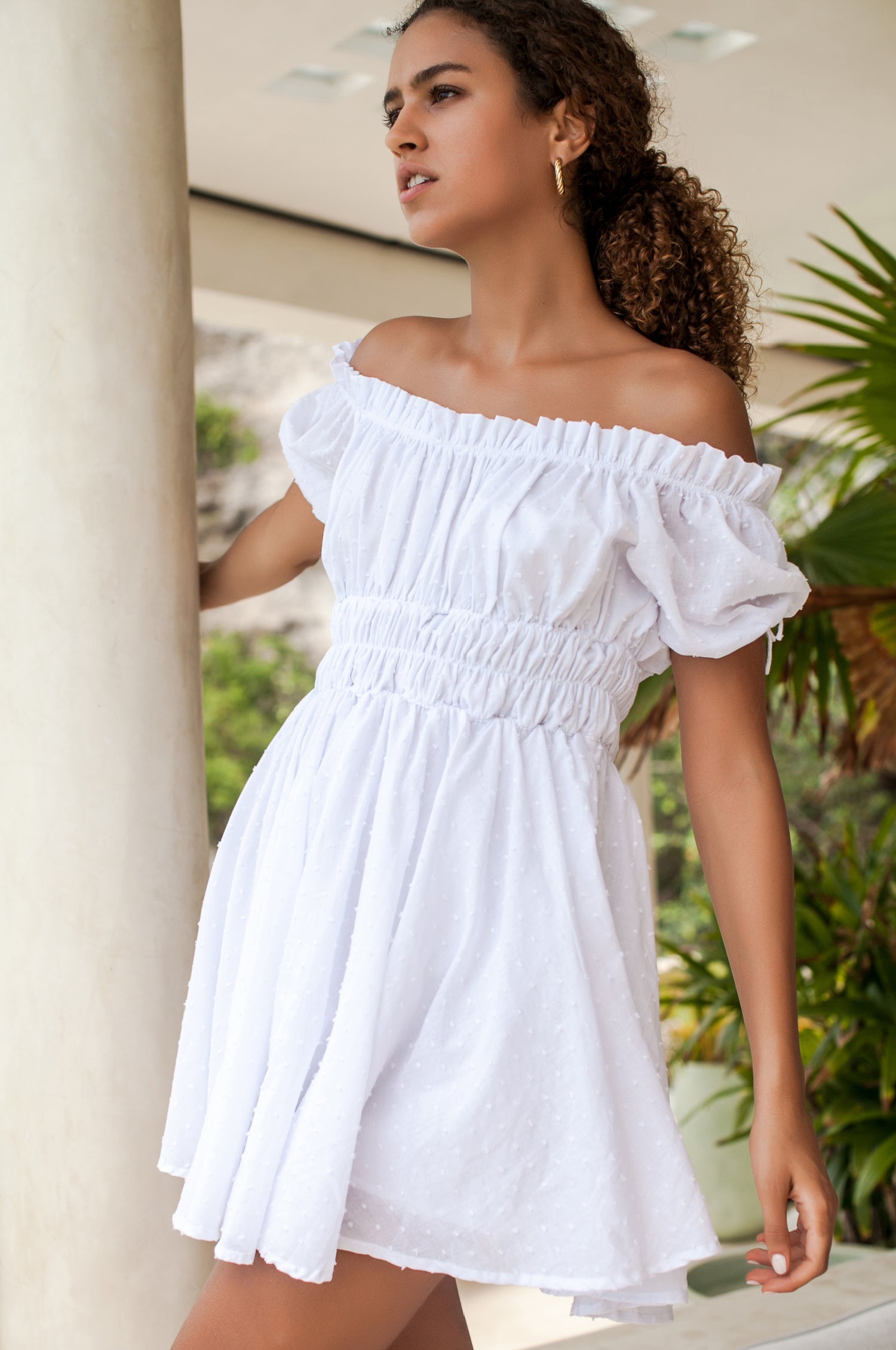 Woman wearing a white off-shoulder cotton mini dress in an outdoor setting with plants and a column.