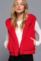 Front close-up of a woman wearing a bright red plush hooded vest with zipper open over a white ribbed long-sleeve top