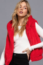 Medium close-up of a woman wearing a red hooded plush zip vest layered over a white ribbed long-sleeve top.