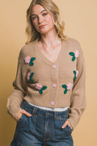 Front close-up view of a khaki cropped cardigan with pink 3D floral appliqués and pink buttons, styled with denim cargo pants.