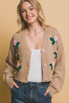 Close-up of open buttoned khaki cropped cardigan with floral appliqués, layered over a white tank top and styled with denim pants.