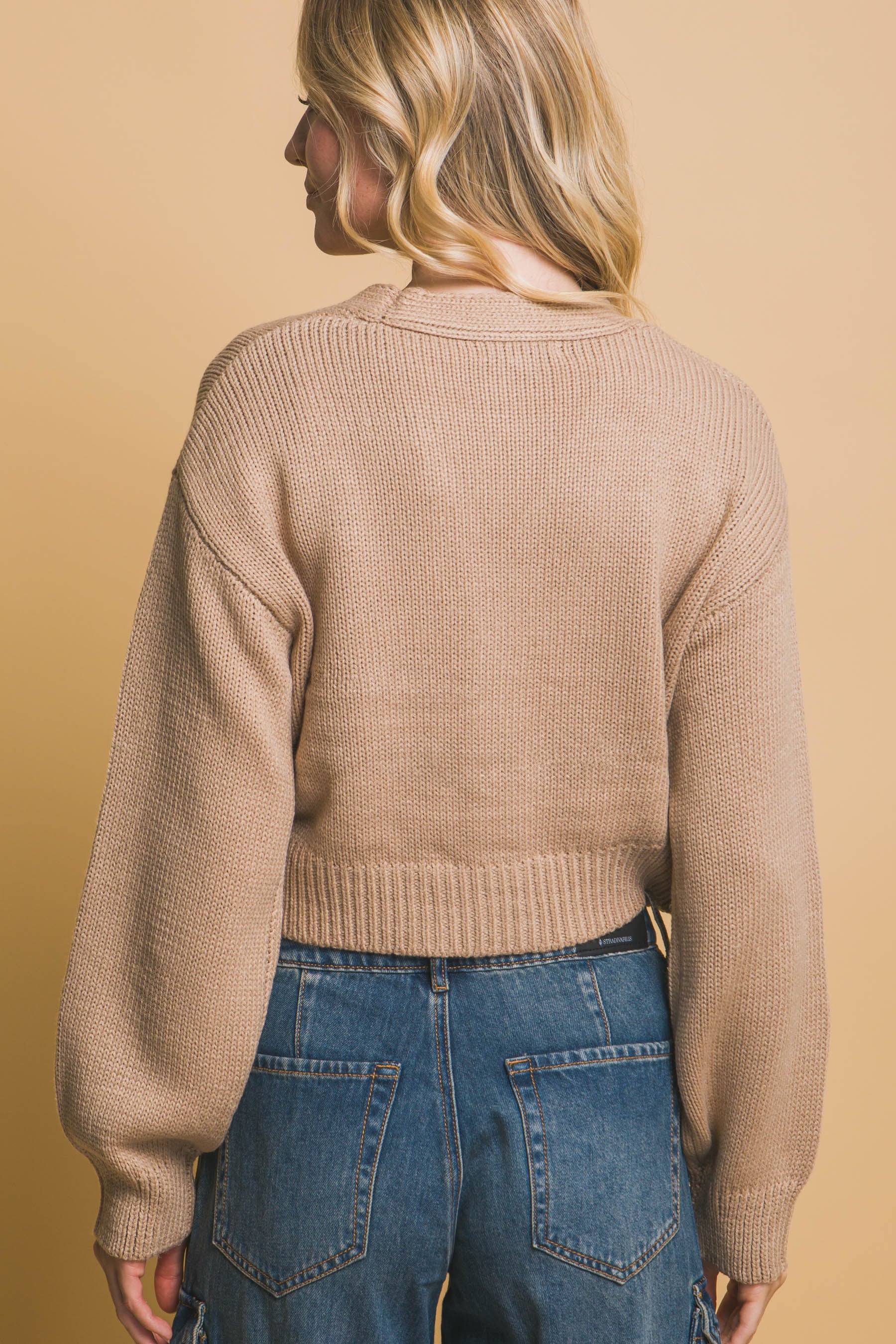 Back view of a khaki cropped cardigan with ribbed hem and long sleeves, shown with denim cargo pants.