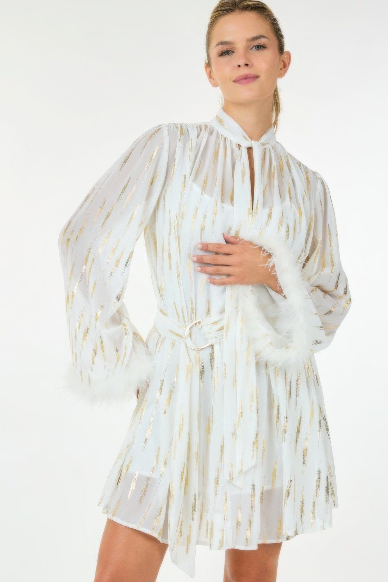 Model wearing white mini dress with gold metallic streaks, tie-neck detail, and feather-trim cuffs, front view close-up.