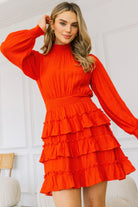 Model wearing a red dress with long sleeves and ruffled layers against a white wall.