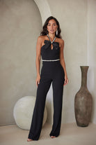 Model wearing a black halter jumpsuit with a satin bow and rhinestone neckline detail, standing in a neutral-toned room with wide-leg pants.