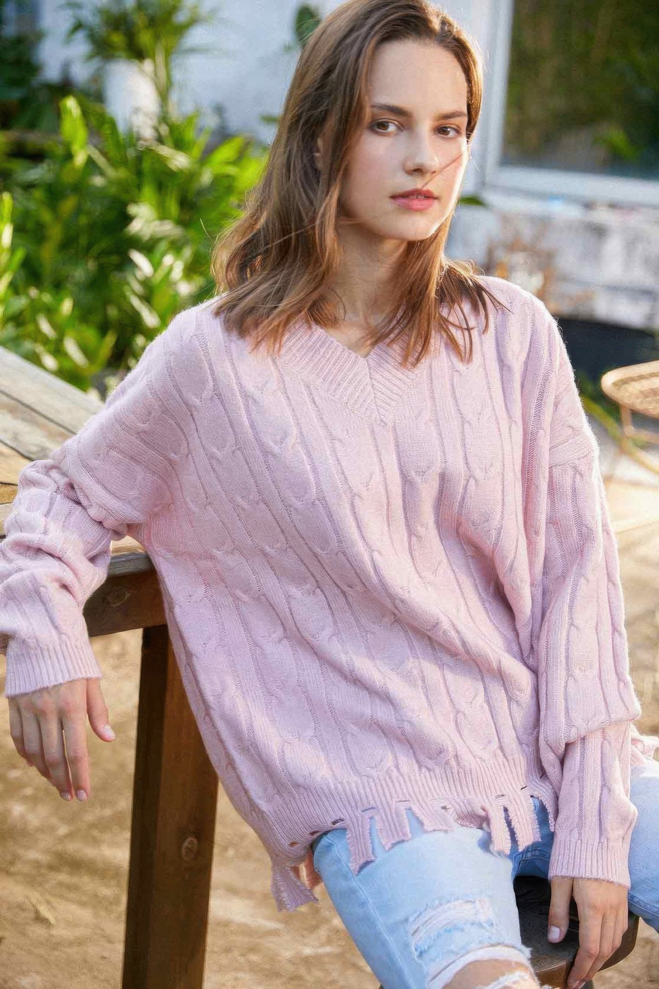 Model sitting outdoors in blush cable knit sweater, leaning on wooden table, showing V-neck and fringe hem-AVAH