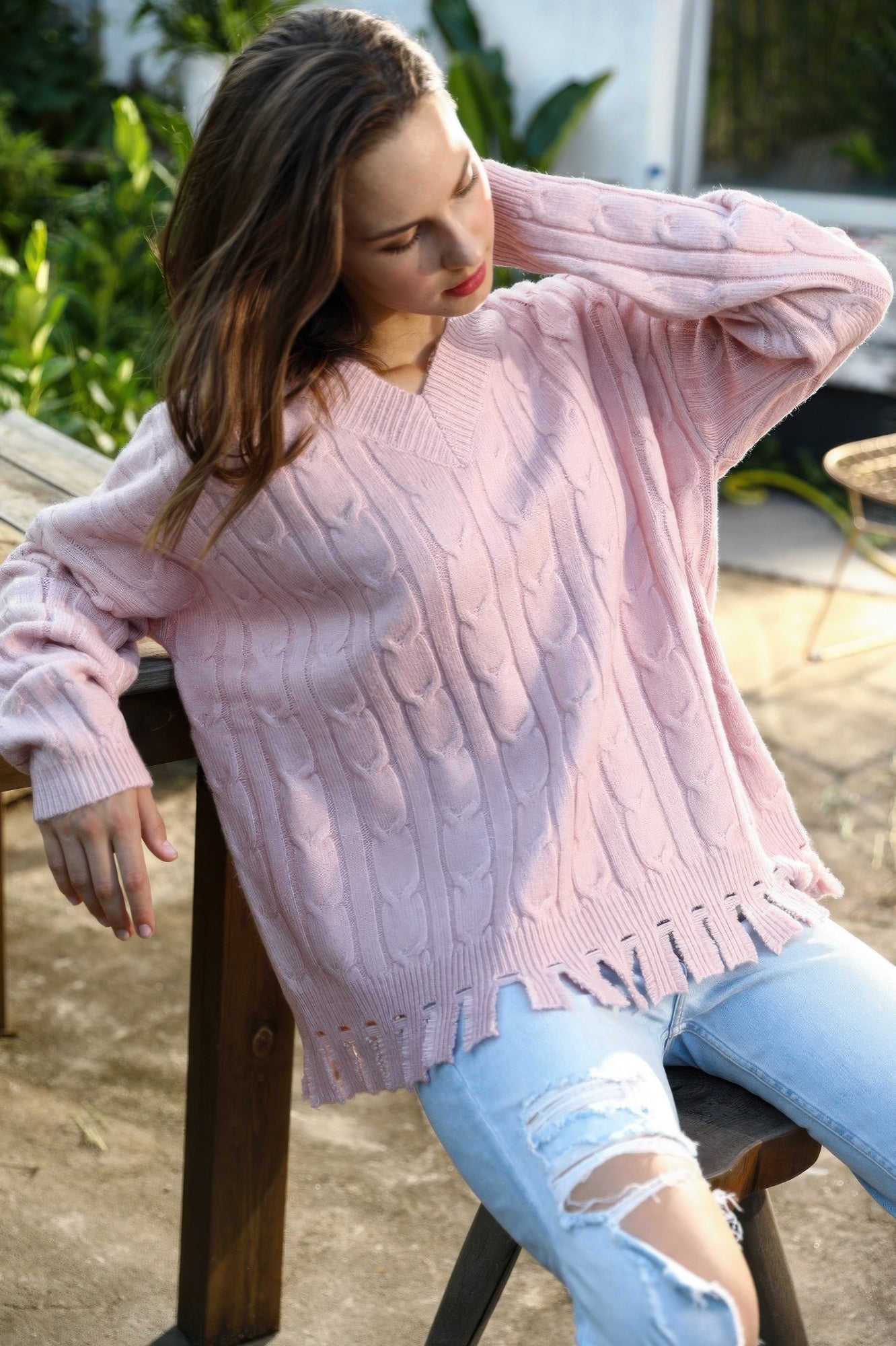 Close-up of model adjusting hair while wearing blush fringe-hem cable knit sweater outdoors.
