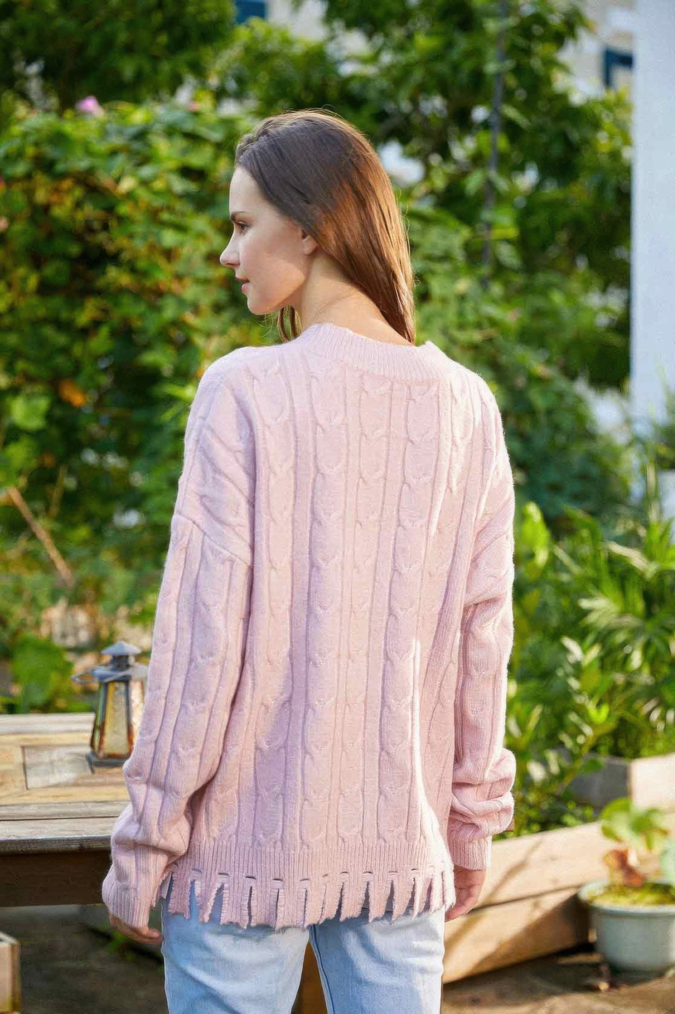 Back view of blush cable knit sweater with fringe hem, worn by model standing outdoors.