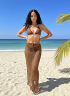 Model wearing a cocoa brown crochet fishnet maxi swim cover up skirt over a brown bikini on a tropical beach.