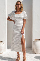 Model wearing the Cala Luna Puff Sleeve Midi Dress in white polka dot while standing in a sunlit courtyard, featuring a scoop neckline, puff sleeves, and side slit.