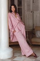 Woman in a pink wide leg pants and matching long sleeve blouse standing in a room with wooden furniture and a white column.