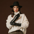 Woman wearing a black hat, white sweater, and black leather gloves against a brown background