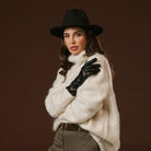 Woman wearing a black hat, white sweater, and black gloves against a brown background