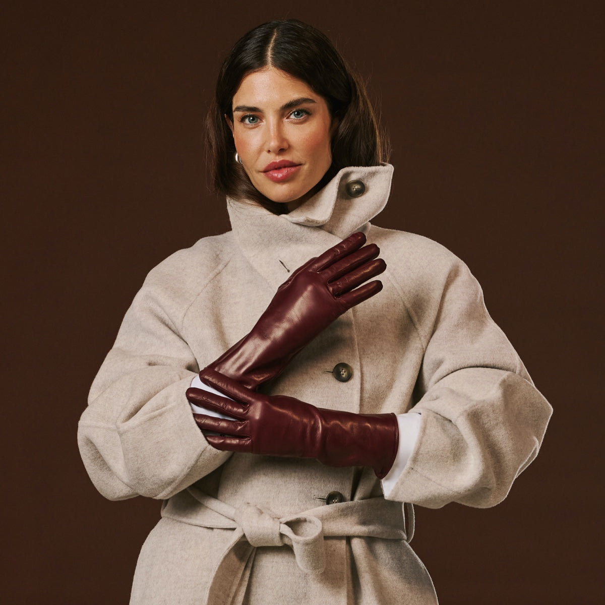 Woman wearing a beige coat and burgundy leather gloves against a brown background