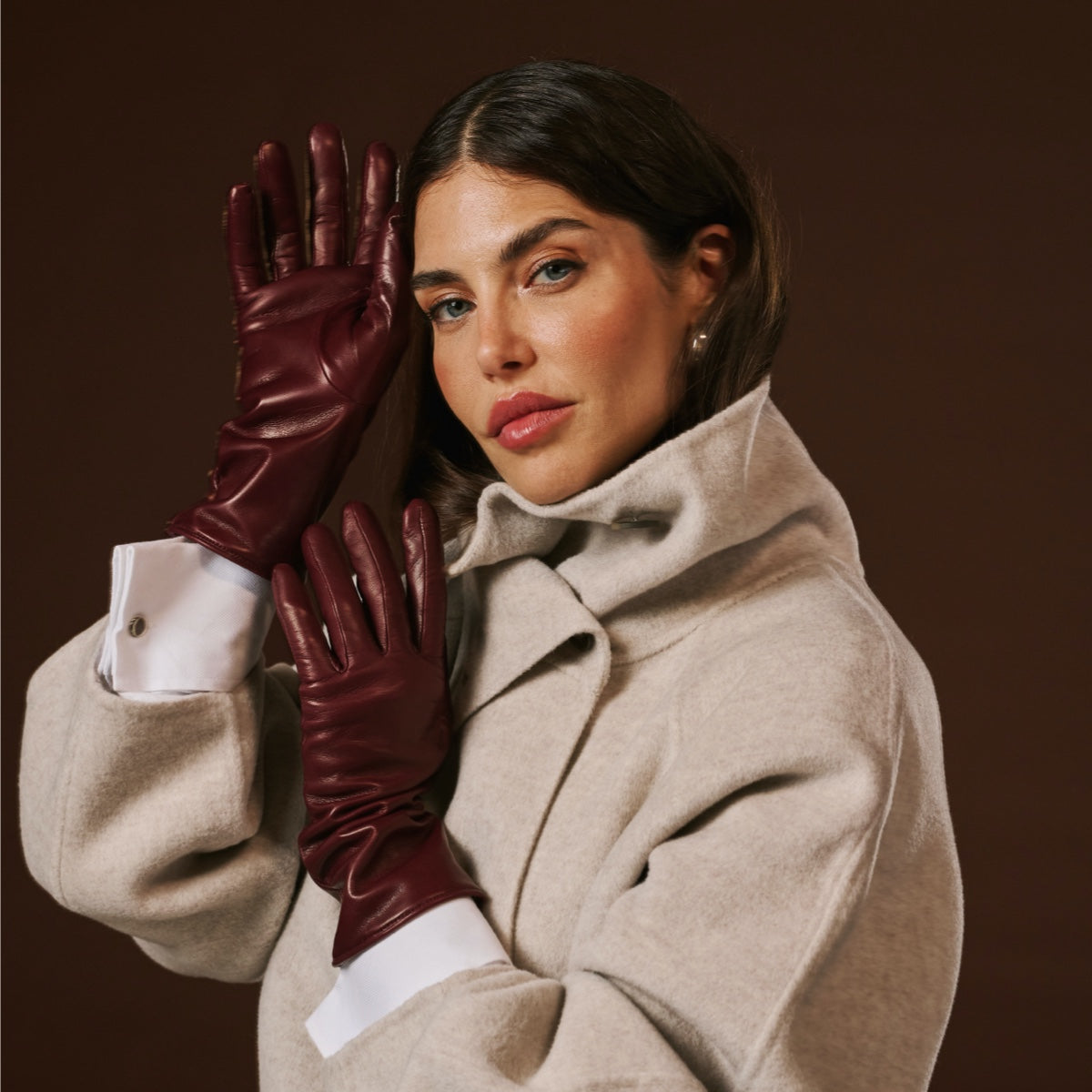 Woman wearing a beige coat and maroon leather gloves against a brown background
