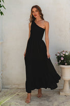 Woman wearing a black one-shoulder maxi dress standing against a light-colored wall with a decorative plant and vase, front view