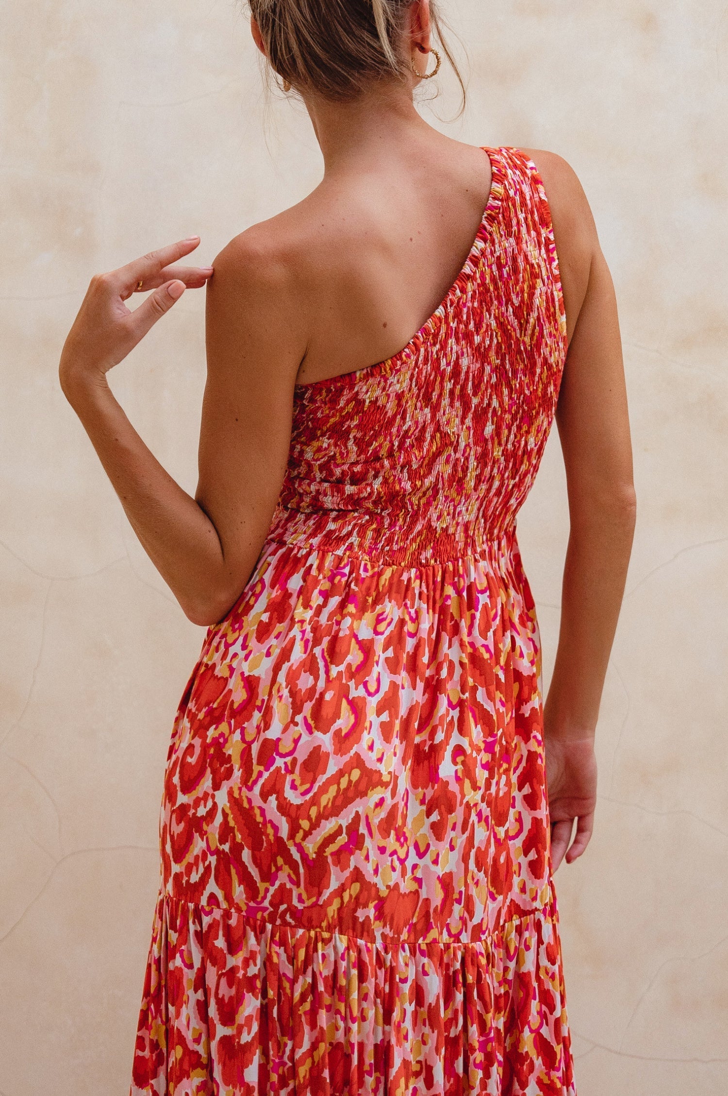 Woman wearing a red and orange patterned one-shoulder maxi dress against a beige wall, back view closeup