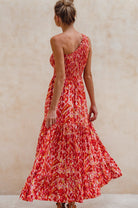 Woman wearing a red patterned one=shoulder maxi dress against a beige wall, back view