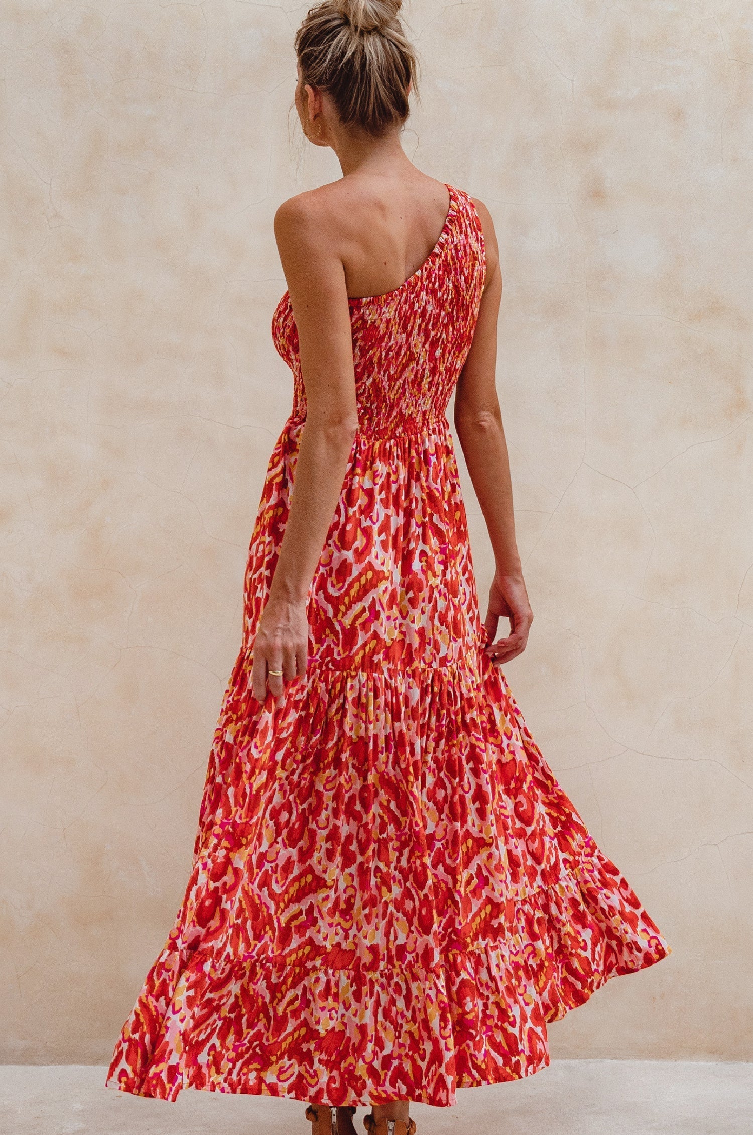Woman wearing a red patterned one=shoulder maxi dress against a beige wall, back view