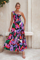 Woman wearing a colorful one-shoulder maxi dress against a neutral wall, front view