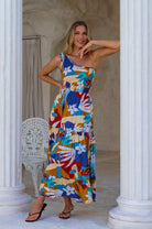 Woman wearing a colorful floral one-shoulder maxi dress standing in a sunlit room with columns, front view