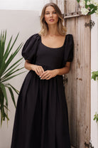 Woman wearing a black midi dress with round neckline and puff sleeves standing against a wooden wall with plants, front view