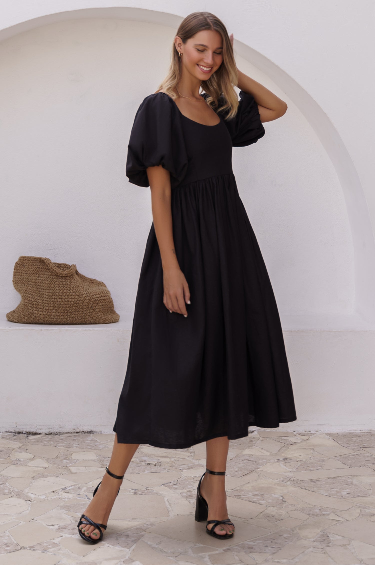 Woman wearing a black midi dress with round neckline and puff sleeves standing against a white wall with a textured surface