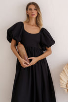Woman wearing a black midi dress with round neckline and puff sleeves standing against a white wall 