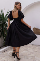 Woman wearing a black midi dress with round neckline and puff sleeves in an outdoor setting, back view