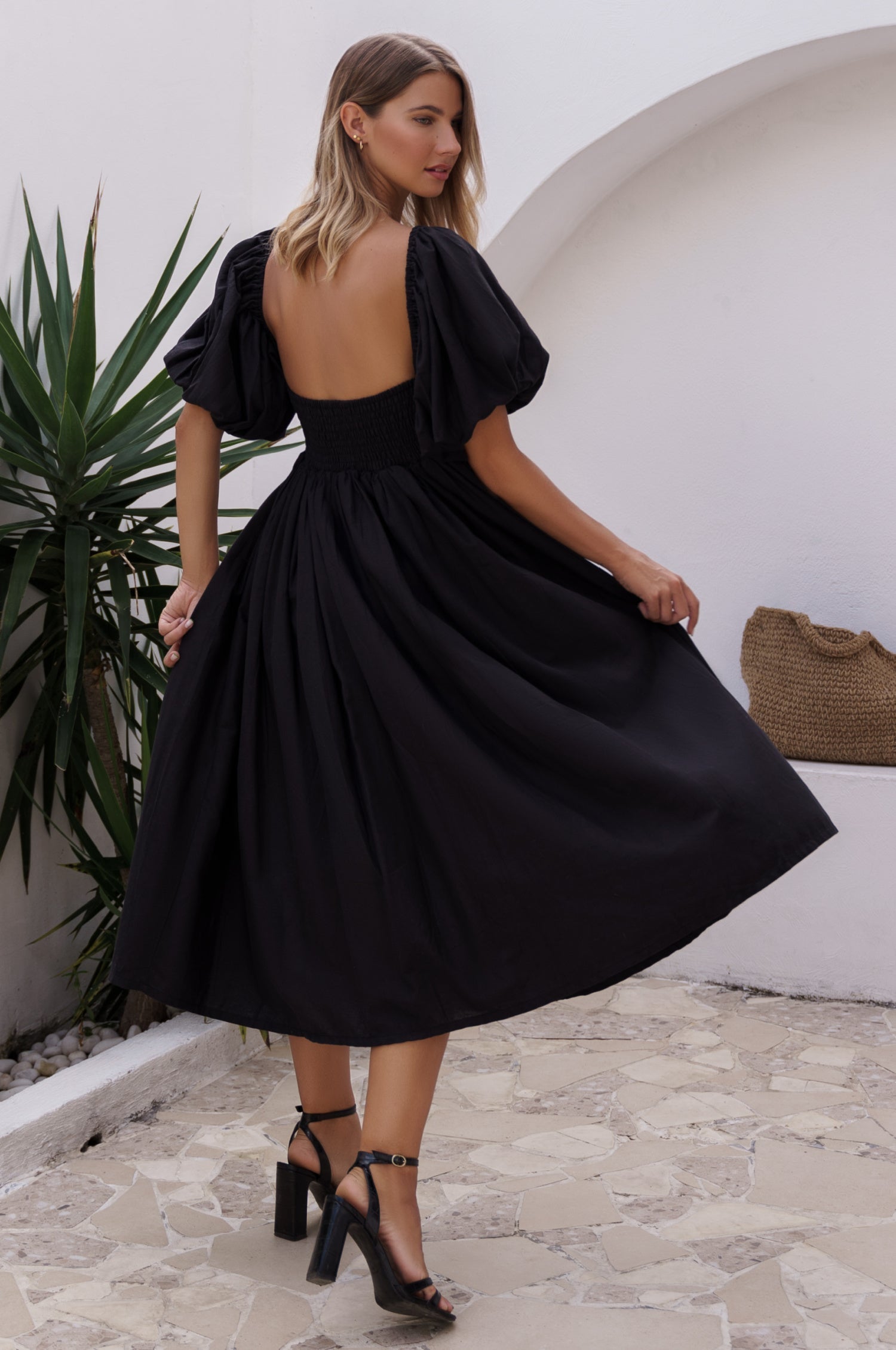 Woman wearing a black midi dress with round neckline and puff sleeves in an outdoor setting, back view