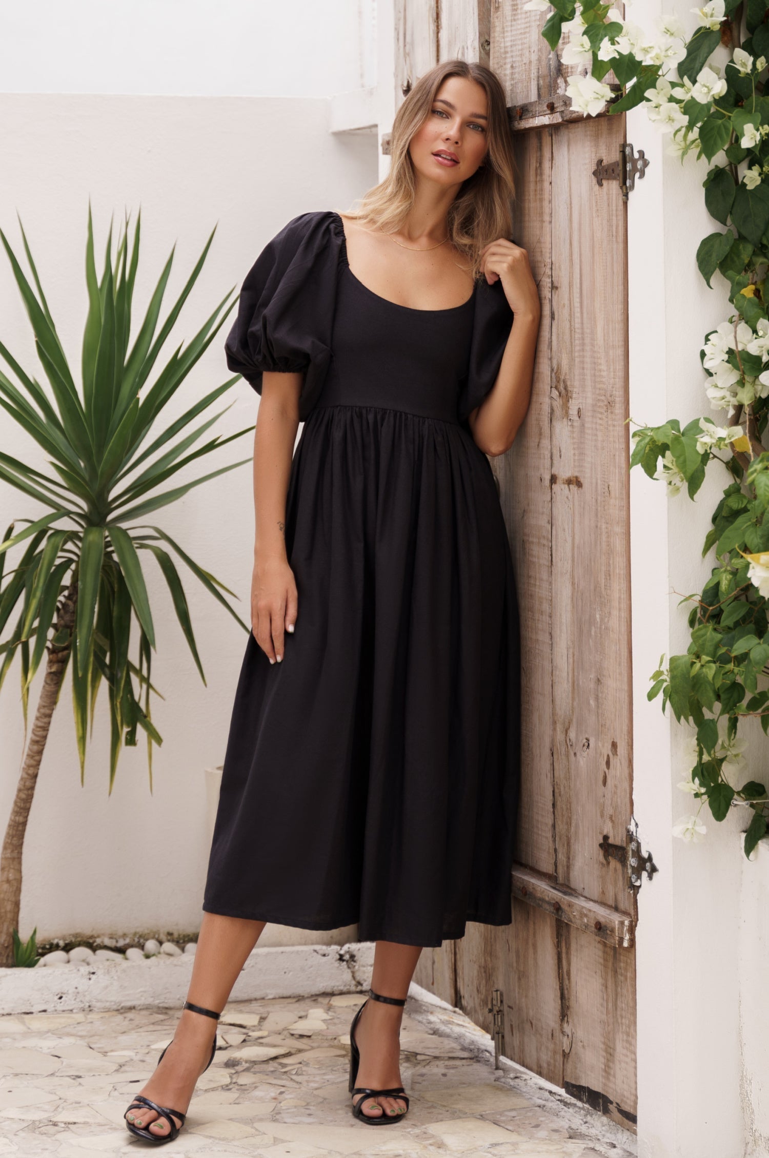 Woman in a black dress midi dress with round neckline and puff sleeves standing in front of a wooden door with plants around.
