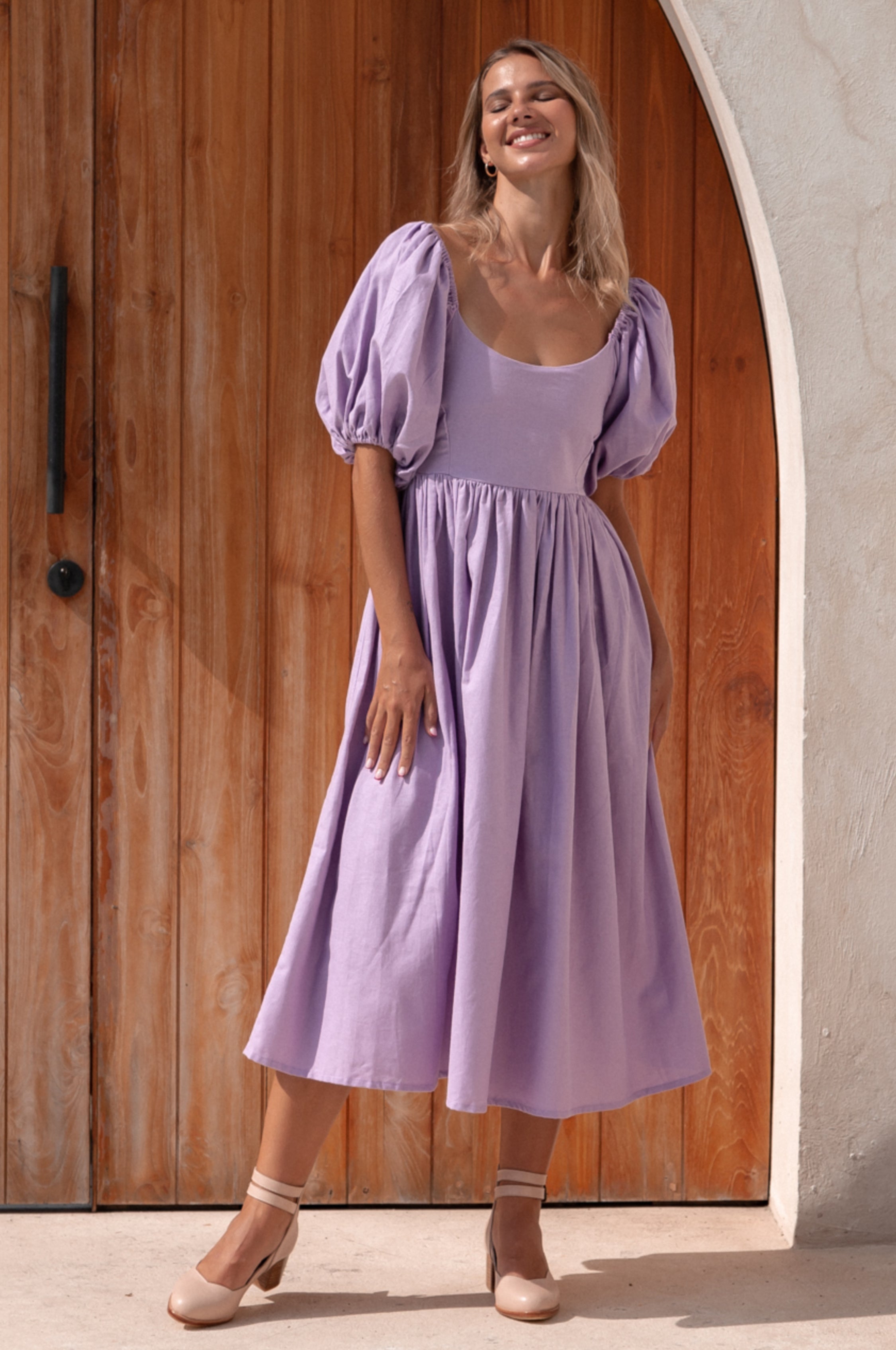 Woman wearing a lavender midi dress with round neckline and puff sleeves standing in front of a wooden door.