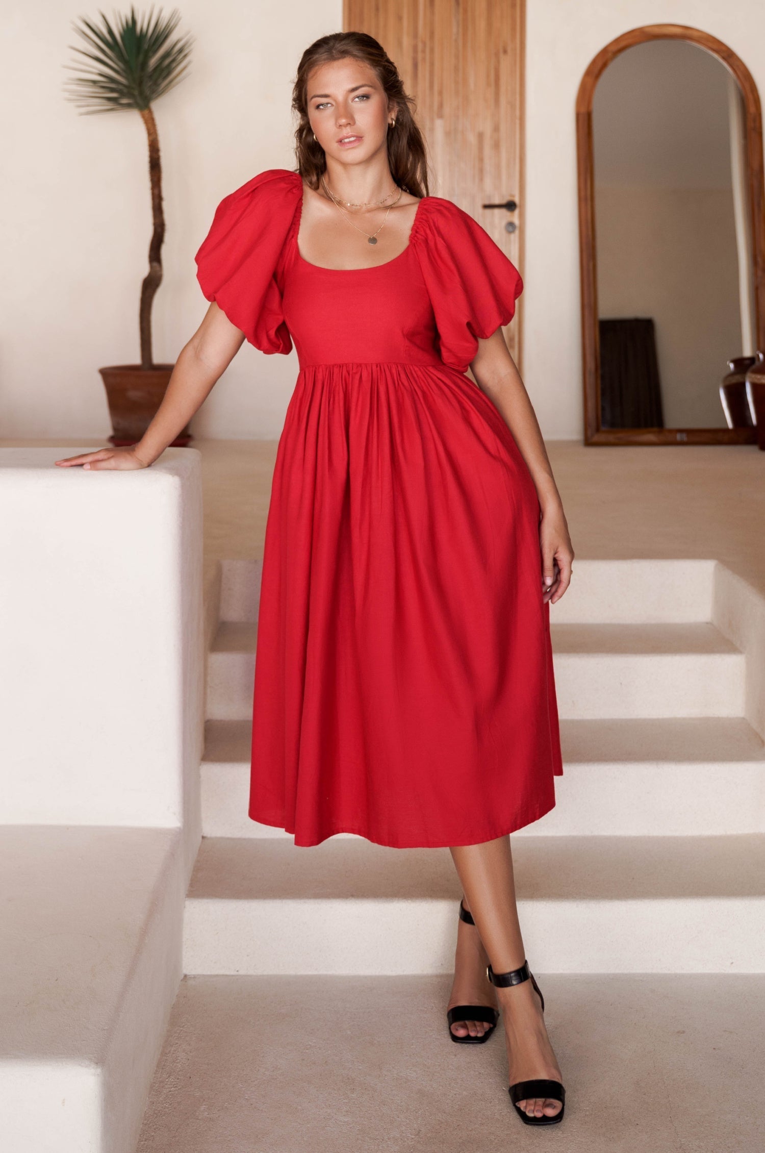 Woman in a red midi dress with round neckline and puff sleeves standing on a staircase in a home setting, front view