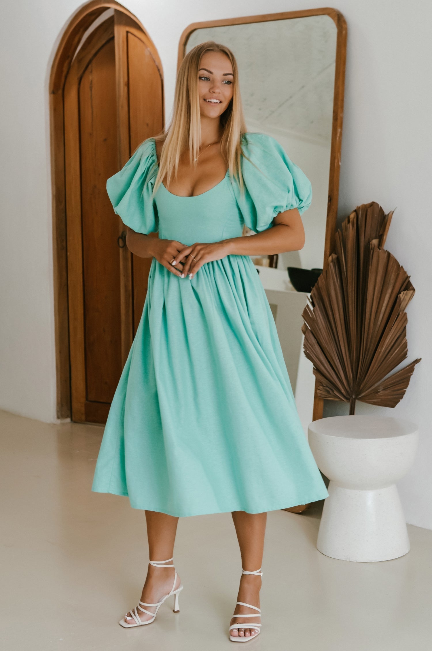 Woman in a turquoise midi dress with round neckline and puff sleeves standing in front of a mirror in a room with a plant and white stool.