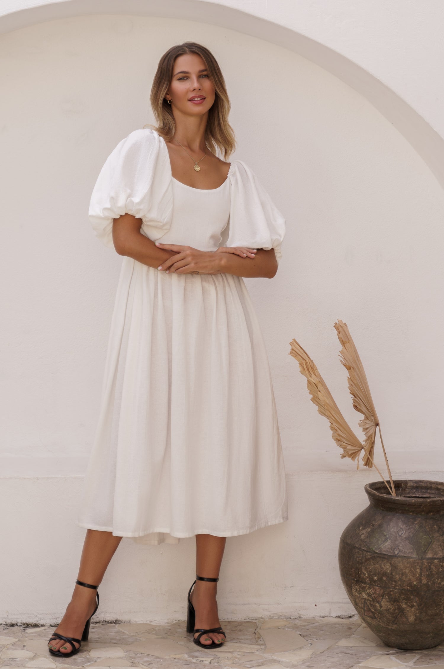 Woman in a white midi dress with round neckline and puff sleeves standing against a white wall with a vase of dried plants, front view