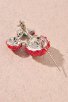 Back view of cherry-shaped brooch covered in red rhinestones with green marquise-cut stones for leaves, set on a silver-tone metal and displayed on a textured beige surface.