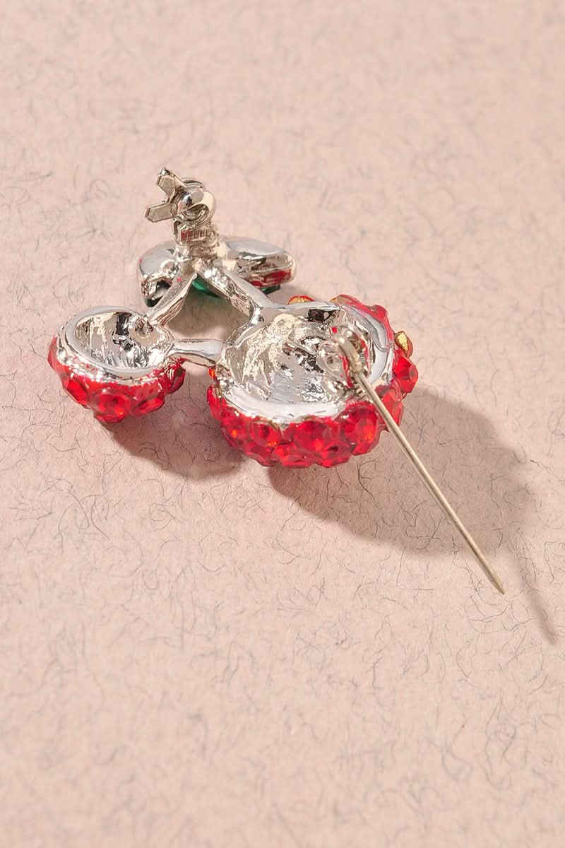 Back view of cherry-shaped brooch covered in red rhinestones with green marquise-cut stones for leaves, set on a silver-tone metal and displayed on a textured beige surface.