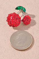 A cherry-shaped brooch covered in red rhinestones with green marquise-cut stones for leaves, set on a silver-tone metal and displayed on a textured beige surface next to a quarter for size comparison.